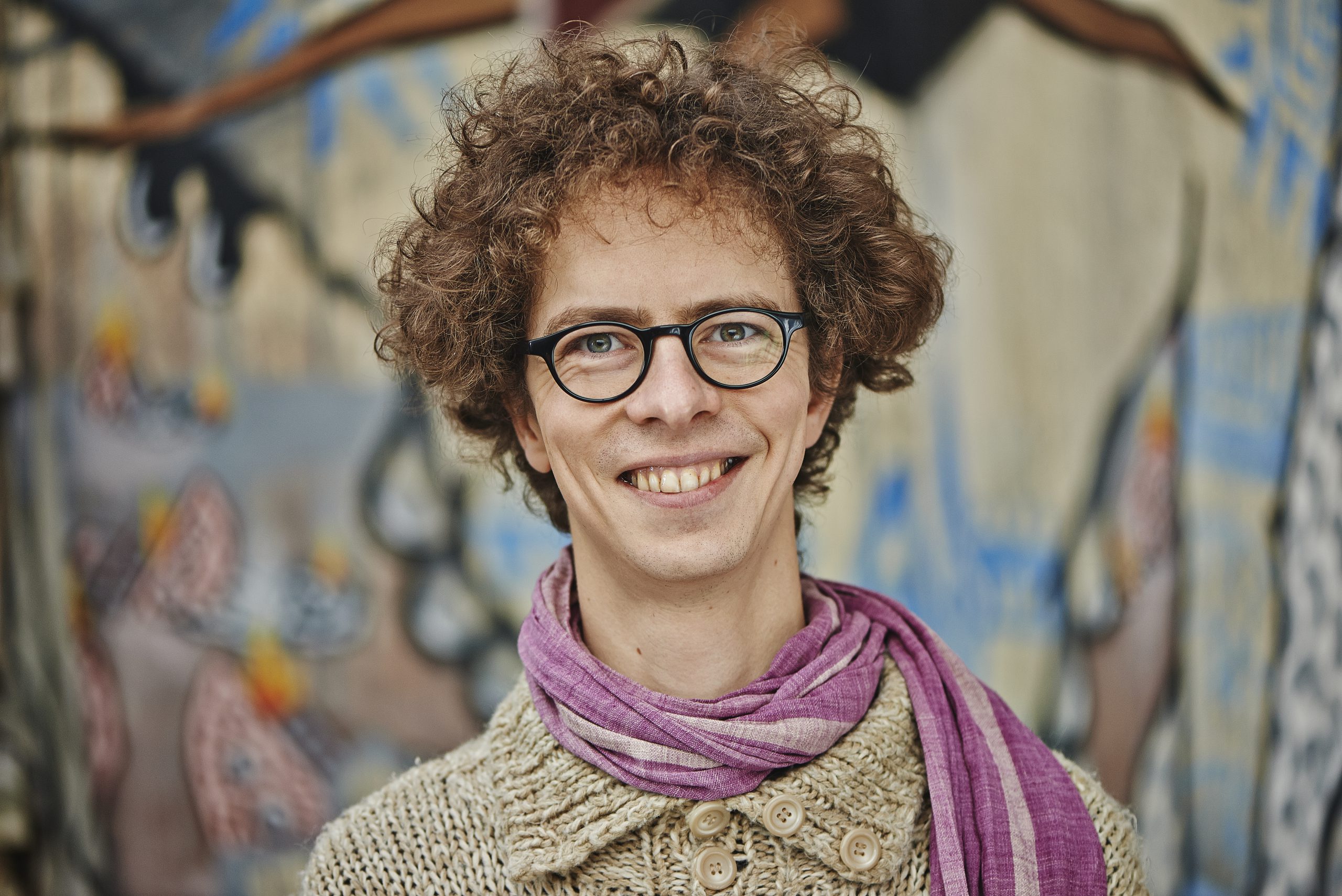 Portraitfoto eines jungen Mannes mit braunen lockigen Haaren. Er schaut lachend in die Kamera.