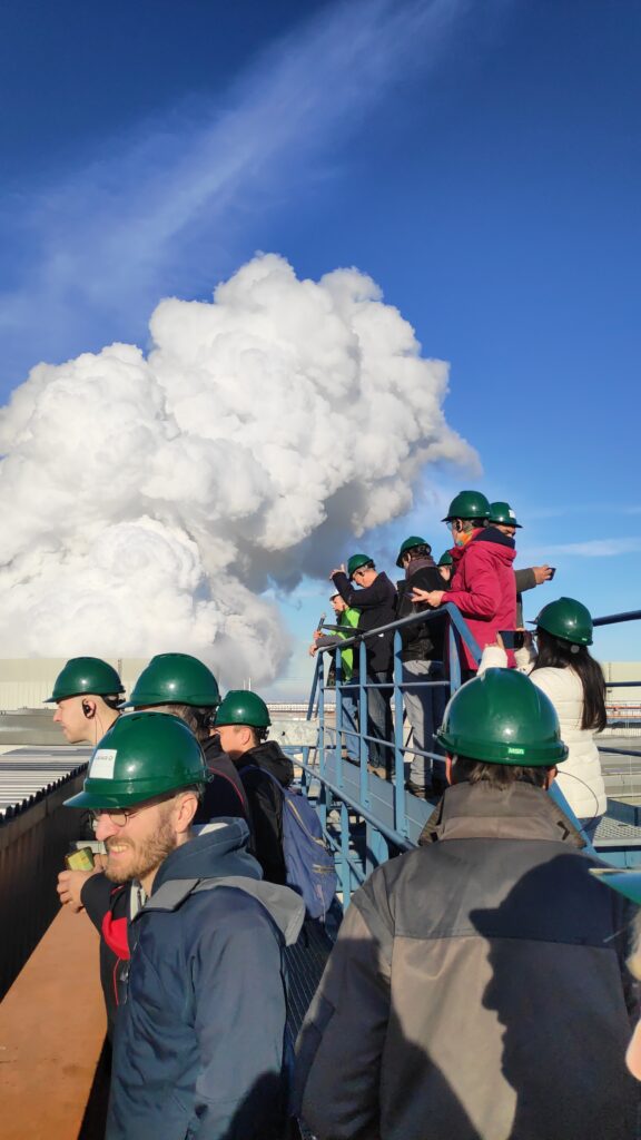 Mehrere Menschen stehen drau&szlig;en auf einem Gel&auml;nder und tragen gr&uuml;ne Schutzhelme. Im Hintergrund sieht man die aufsteigenenden Wasserdampfwolken aus den K&uuml;hlt&uuml;rmen..