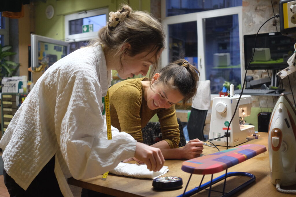 Zwei Personen stehen an einem Tisch mit N&auml;hmaschine und B&uuml;geleisen.