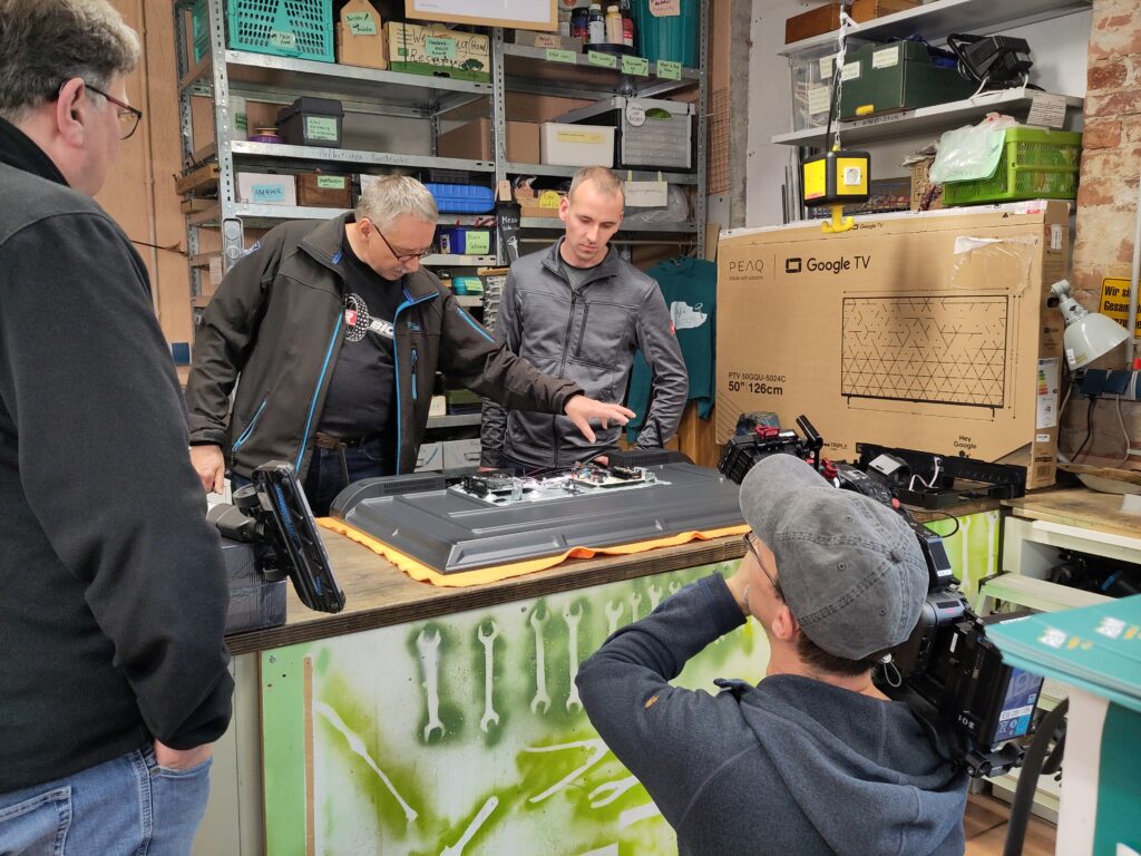 Zwei Personen arbeiten an einem Flachbildschirmfernseher, der auf der Werkbank liegt. Eine Person kniet davor und richtet eine Kamera, die sie auf der Schulter h&auml;lt auf das Geschehen.
