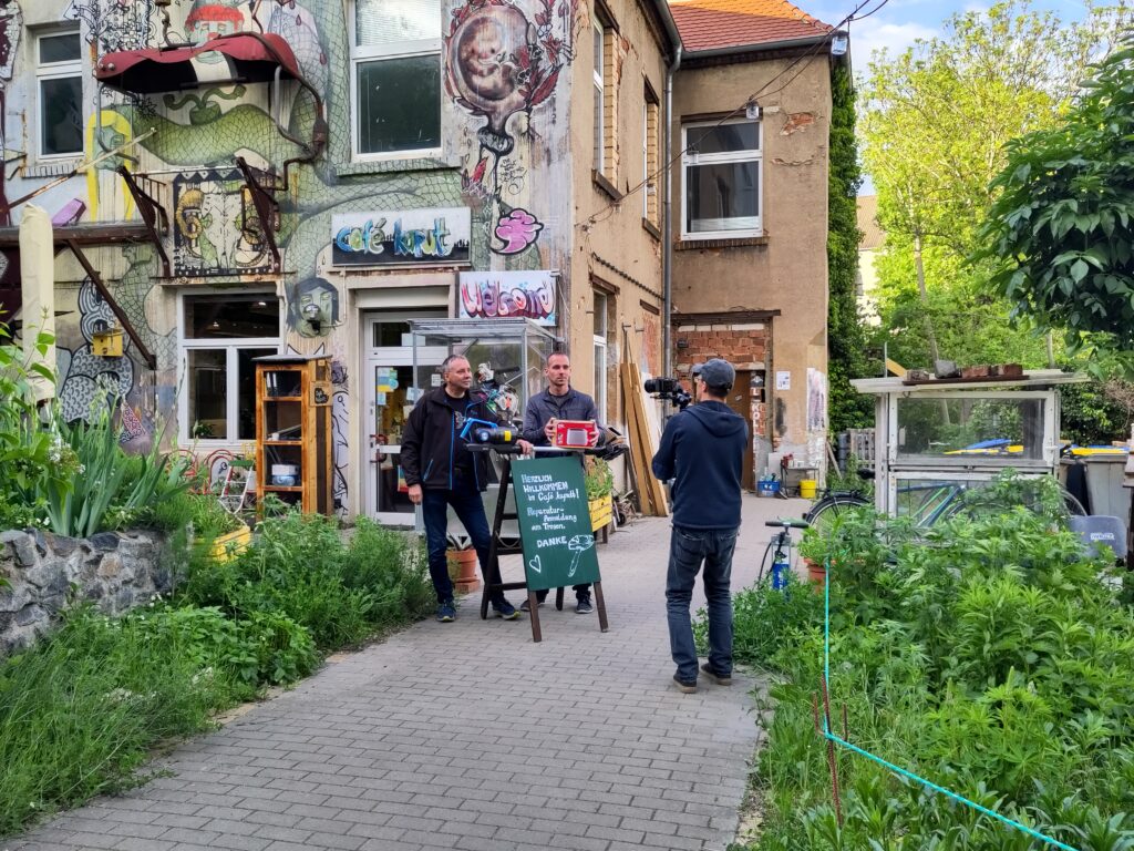 Zwei Personen stehen vor einem Aufsteller vor dem Caf&eacute; kaputt. Ein Mensch mit einer Kamera filmt die zwei.