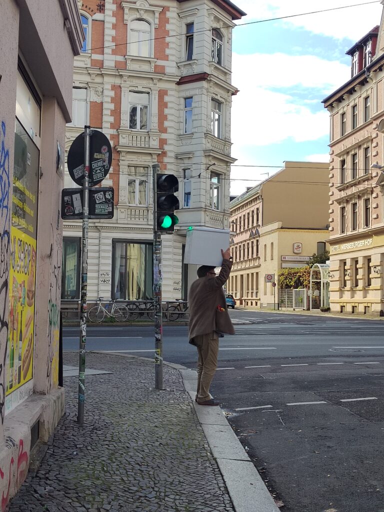 Eine Person tr&auml;gt einen kleinen K&uuml;hlschrank und wartet an einer Ampel.