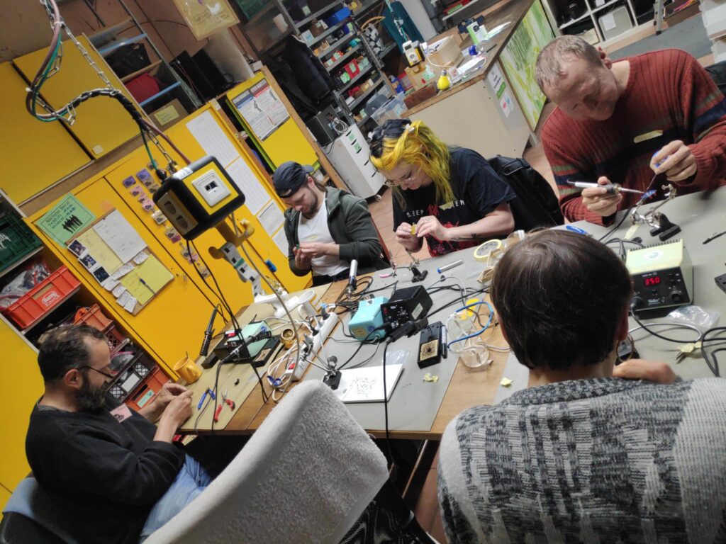 Mehrere Menschen arbeiten gemeinsam an einem Werkstatttisch und l&ouml;ten.