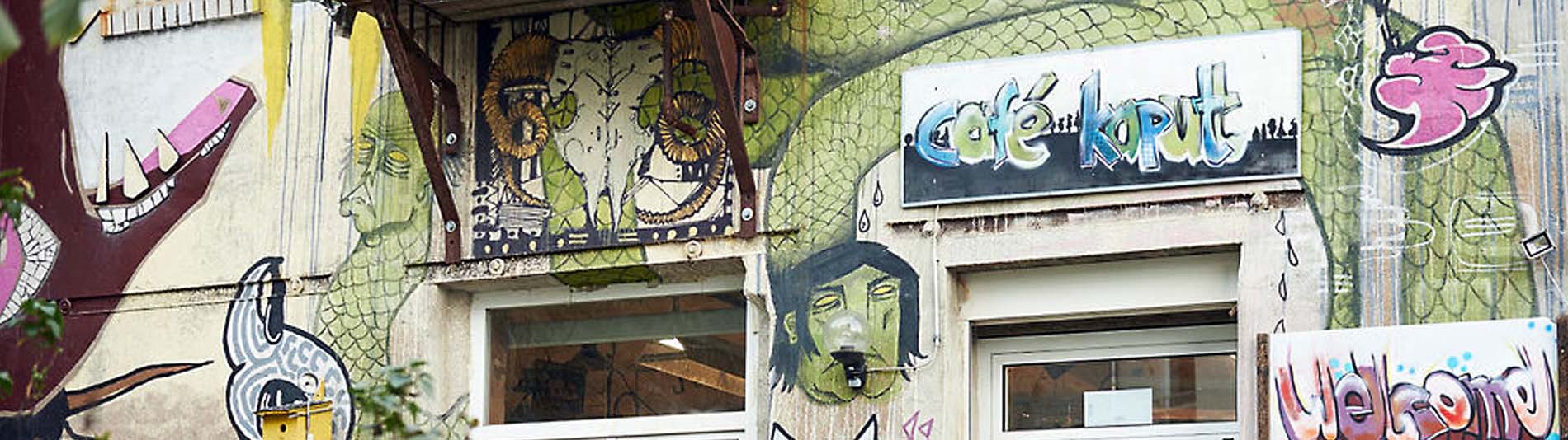 Die bunt bemalte Fassade des Café kaputt ist zu sehen mit den selbst beschriebenen Schildern: Café kaputt und welcome.