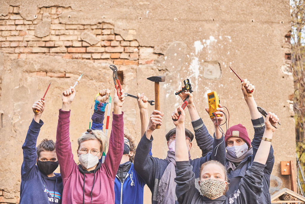 Eine Gruppe an Menschen halten in beiden Händen Werkzeuge hoch. Sie stehen vor einer Mauer und tragen Masken.