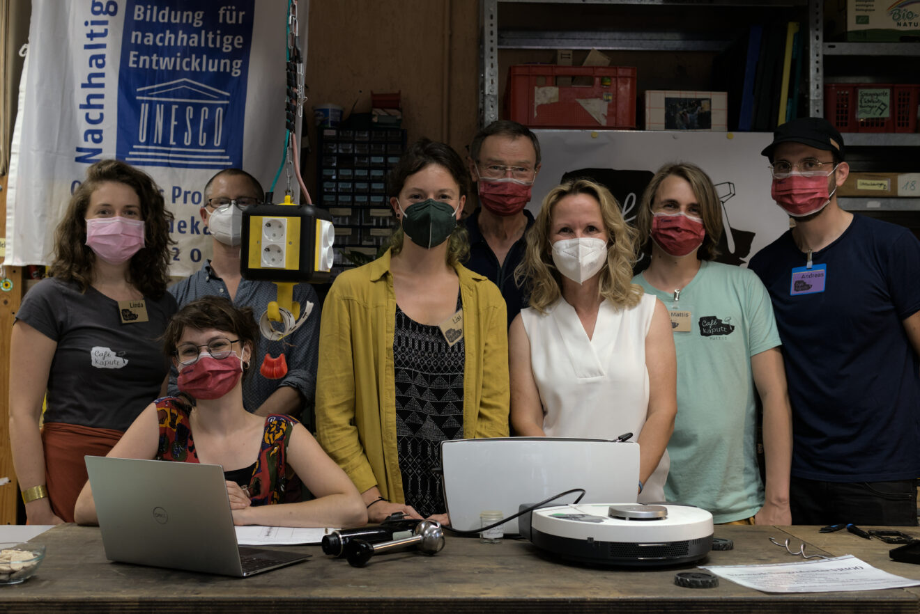 Auf dem Bild stehen acht Personen hinter einer Werkbank. Von Rechts nach Links sind zu sehen: Andreas Blum, Matthias Grill, Steffi Lemke, Eberhard Breuer, Lida Kuhley, Anna Hardock, Tom Hansing, Linda Marie Zebe