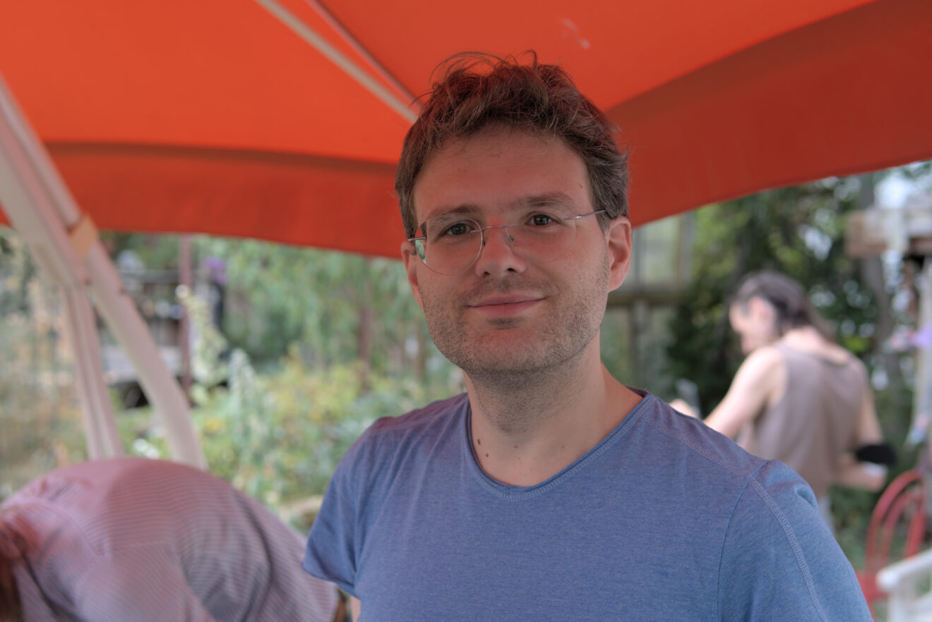 Auf dem Bild ist Gunnar Warnecke zu sehen: Er lächelt in die Kamera. Er steht unter einem roten Schirm und trägt ein blauer T-Shirt.