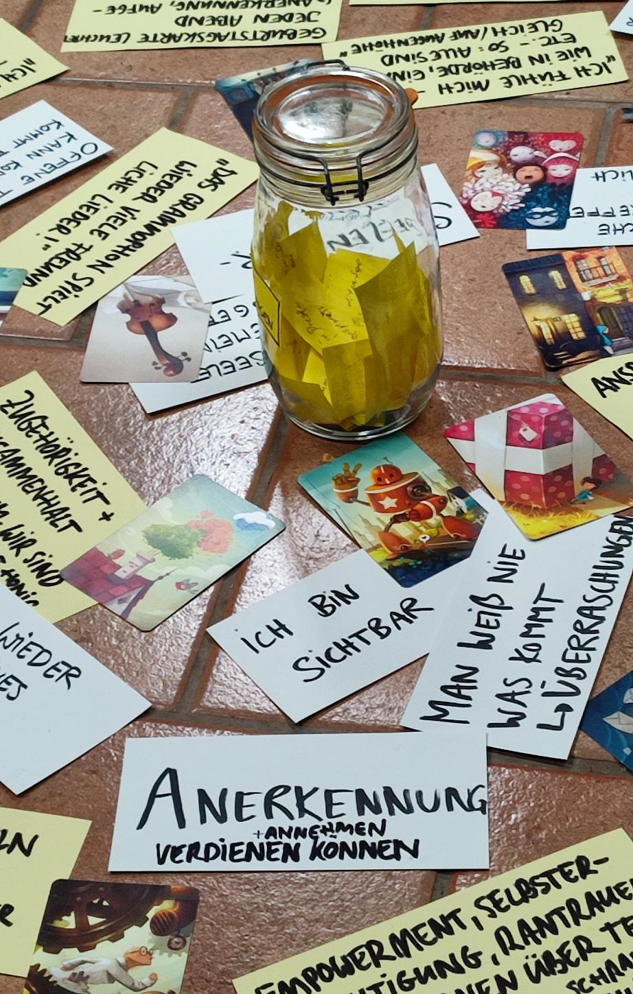 Auf dem Bild steht ein Einmachglas, gefüllt mit gelben Zetteln. Ringsherum liegen beschriftete Zettel und bunte Karten. Alles liegt auf einem Fliesenboden.