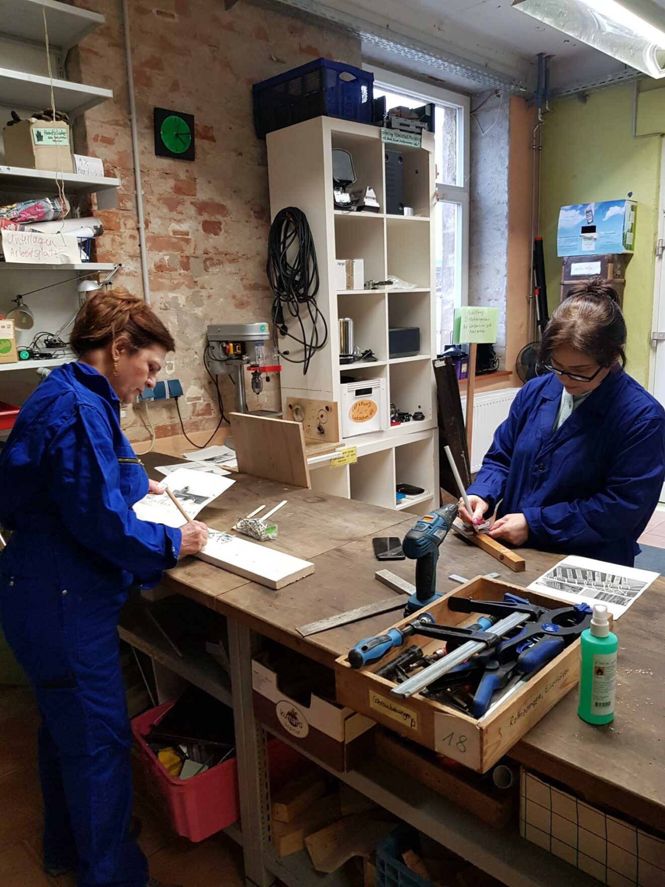 Zwei Frauen in blauen Overals stehen an der Werkbank und arbeiten an ihren Holz-Projekten.