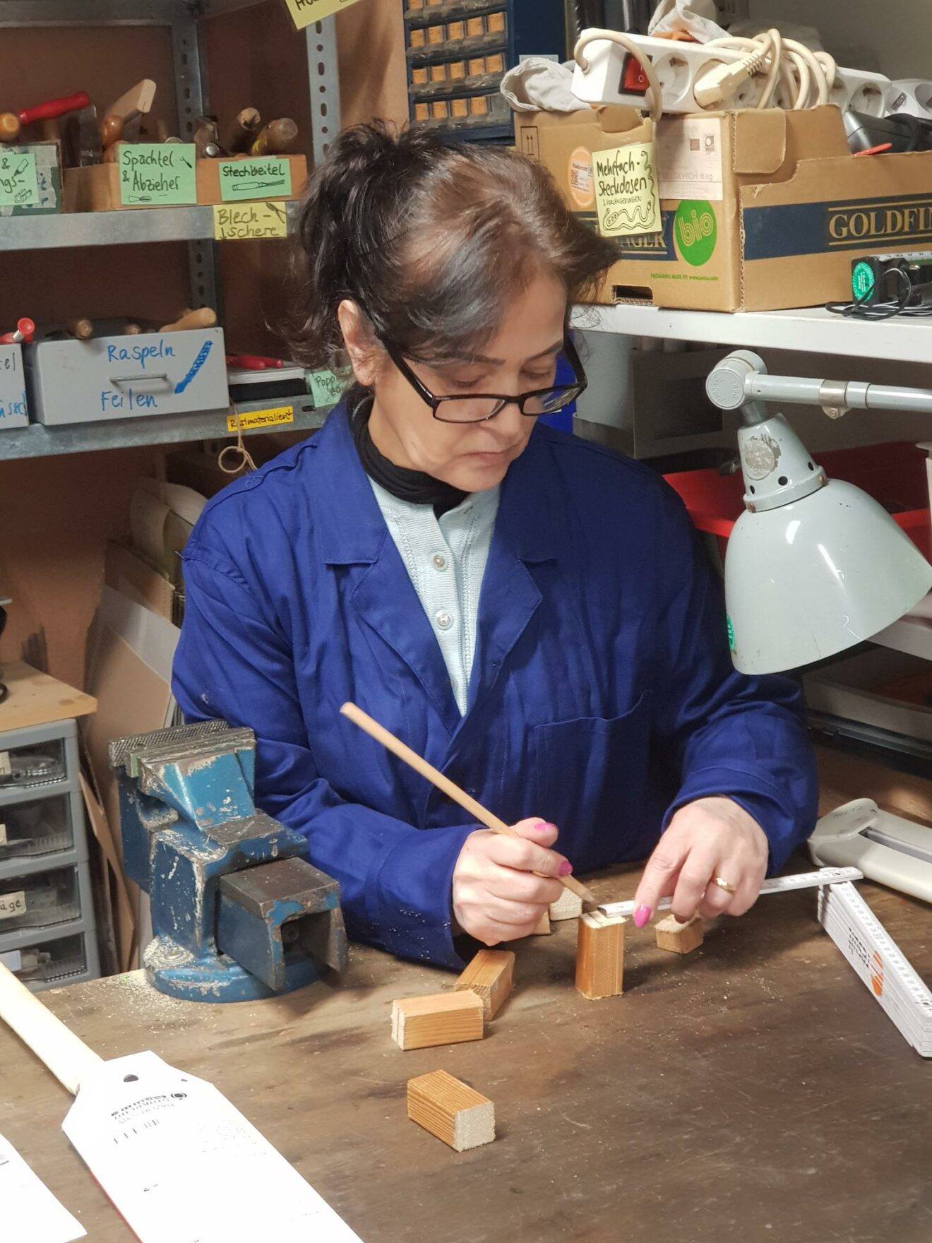 Eine Frau mit Brille steht an der Werkbank und zeichnet mit Bleistift auf ein Stück Holz. Sie trägt auch einen blauen Overall.
