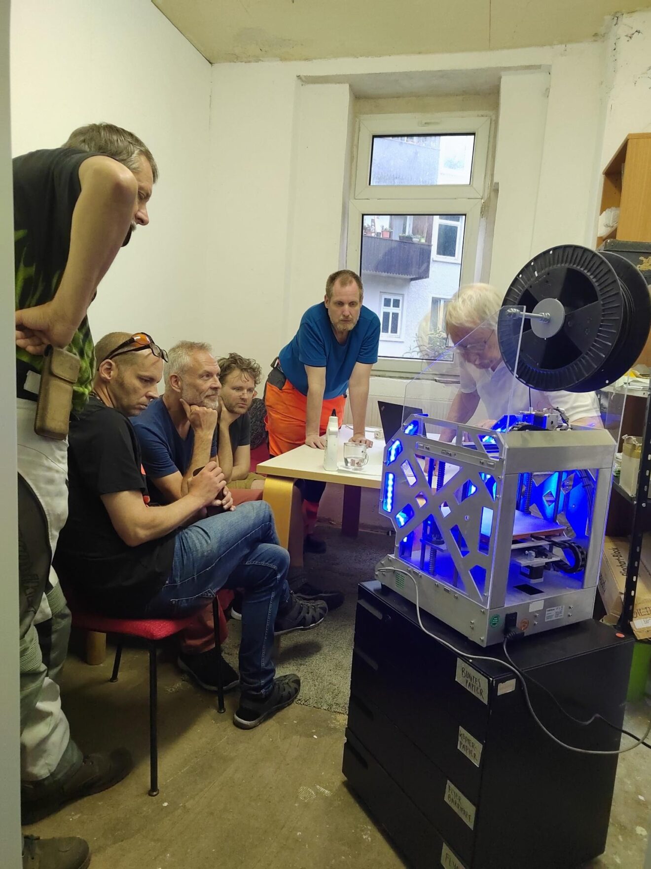 Fünf Menschen sitzen um den 3D-Drucker und schauen beim Drucken zu. Der Drucker ist von innen blau angeleuchtet.