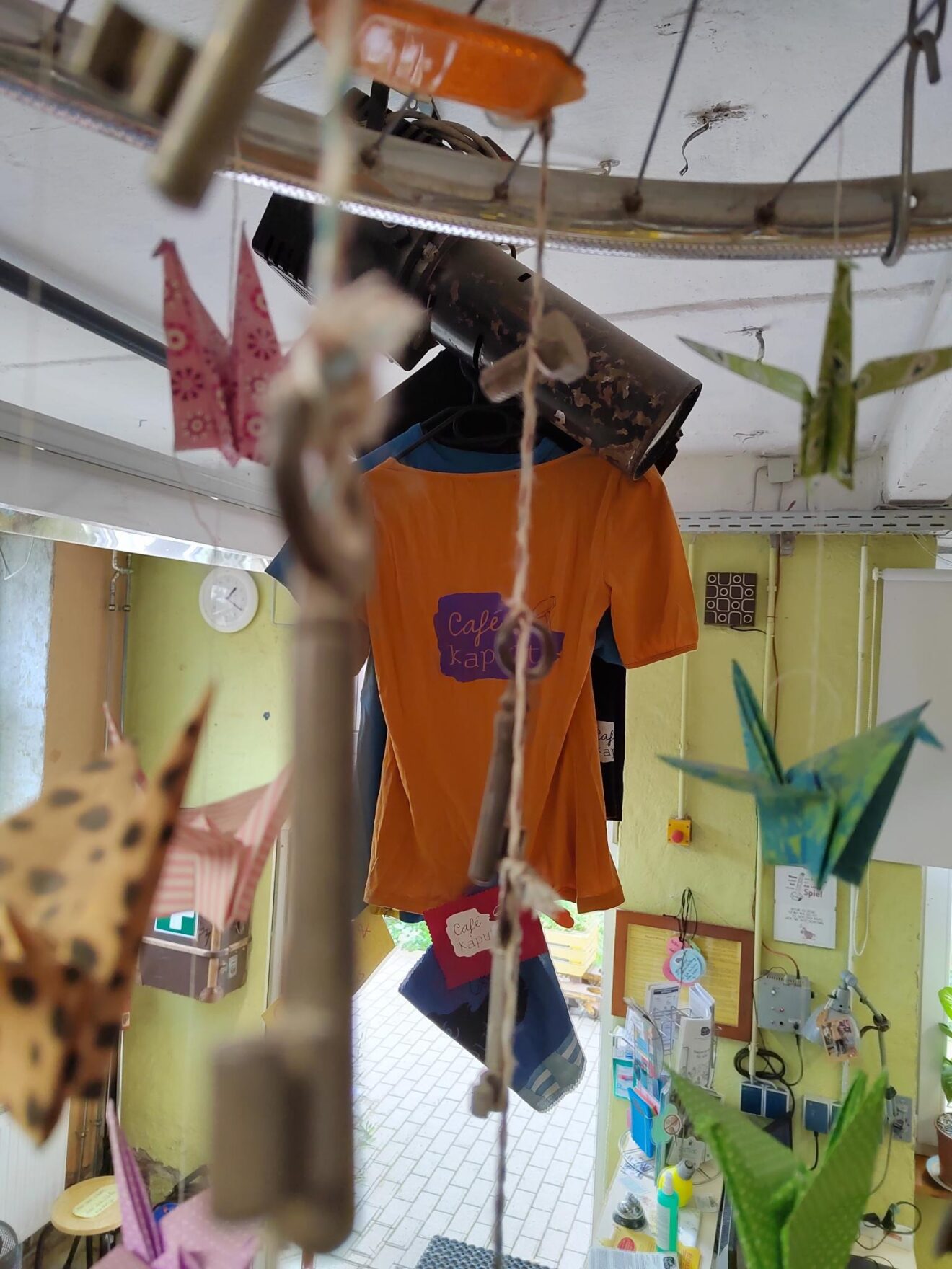 An der Decke des Café kaputt hängen verschiedene T-Shirts mit Café kaputt Logo. Im Vordergrund hängt ein orangefarbenes T-Shirt mit lilafarbenem Café kaputt Logo.