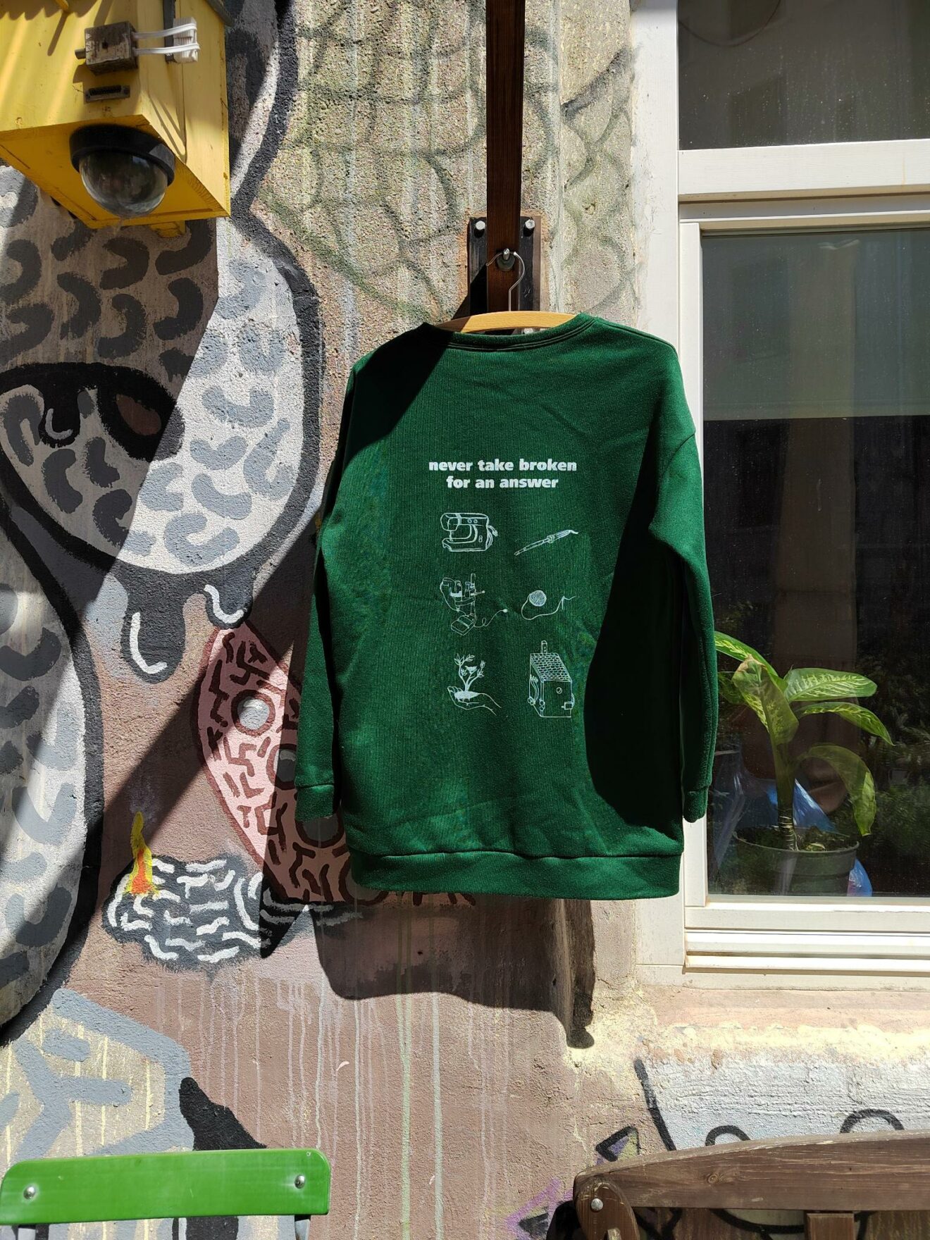 Ein grüner Pullover hängt an der Außenwand vor dem Café kaputt. Darauf sind verschiedene Piktogramme gedruckt und der Spruch "never take broken for an answer".