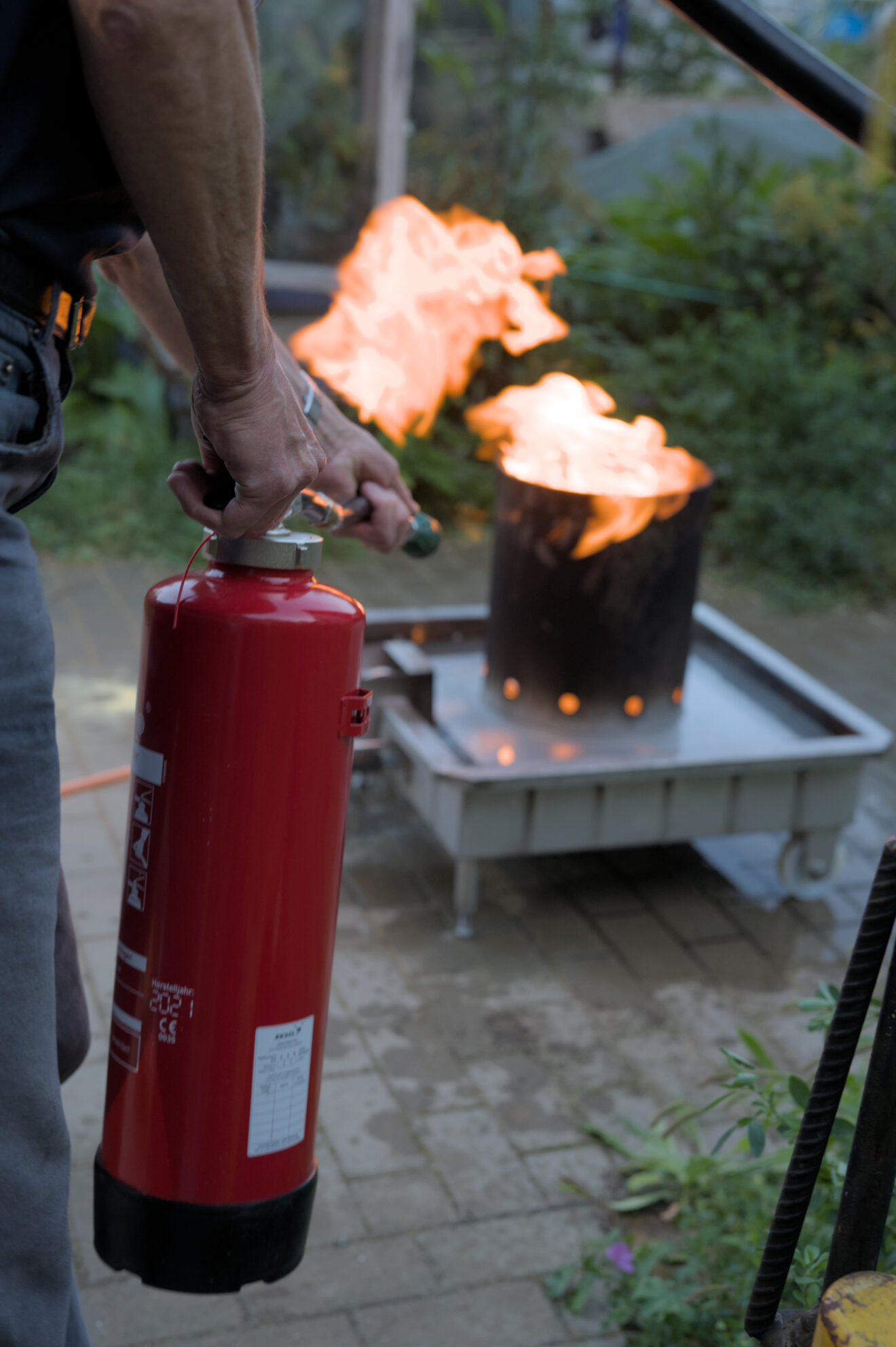 Im Vordergrund sind links im Bild Hände zu sehen, die einen roten Feuerlöscher halten. Im Hintergrund brennt Feuer in einem runden Gefäß. Die Flammen sind orangefarben und steigen auf.