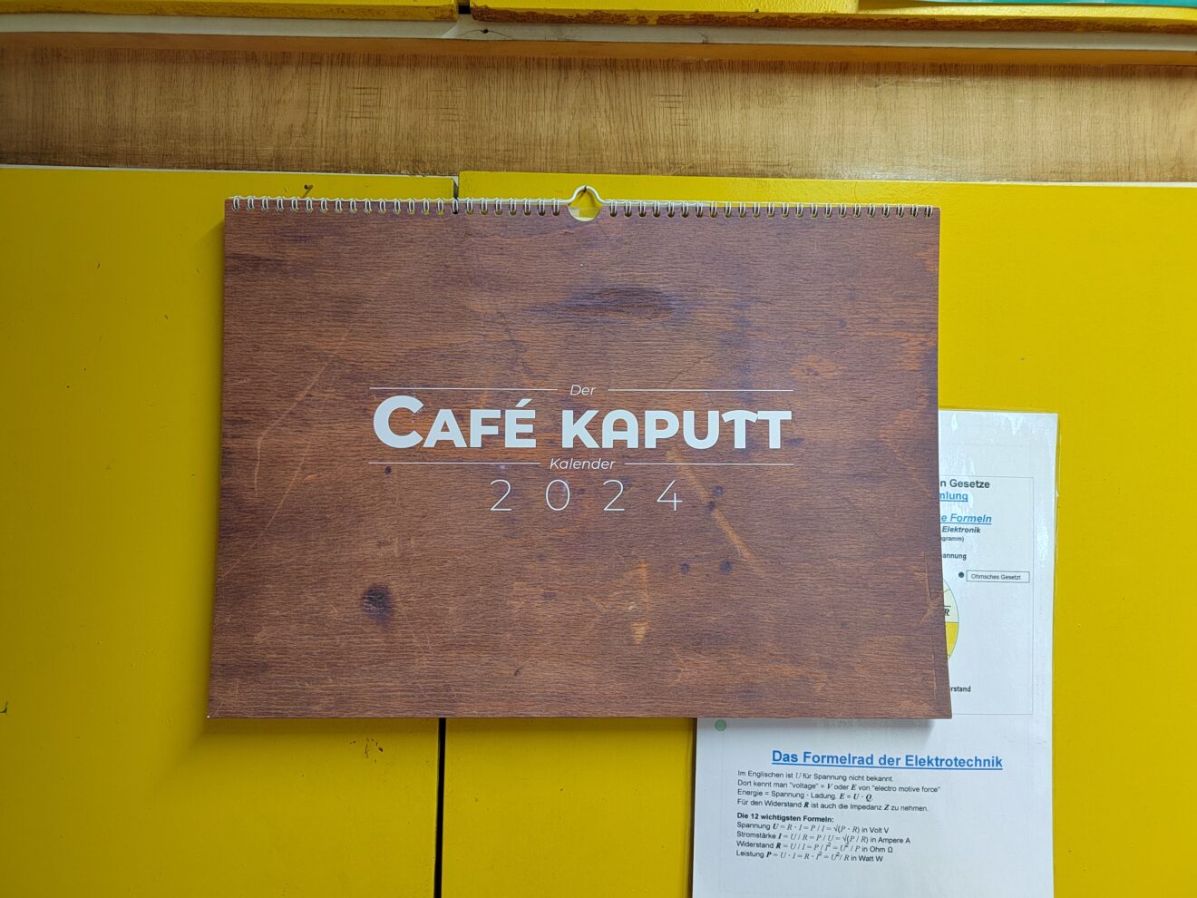 Das Bild zeigt das Deckblatt vom Café kaputt Kalender für 2024. In weißer Schrift steht "Café kaputt 2024" im Hintergrund ist eine Nahaufnahme vom Holz der Werkbank im Café kaputt zu sehen. 