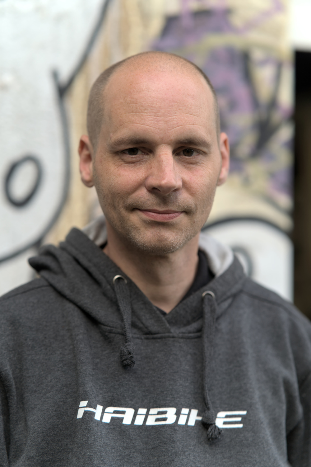 Emma lächelt in die Kamera. Er trgt einen grauen Kapuzenpullover mit einem weißen Schriftzug. Der Hintergrund ist verschwommen.