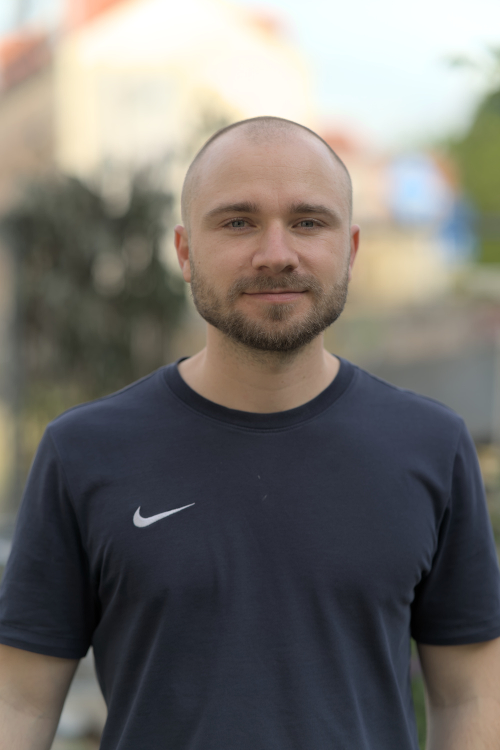 Manu lächelt in die Kamera. Er trägt ein dunkelblaues T-shirt und einen Dreitagebart. Der Hintergrund ist verschwommen.