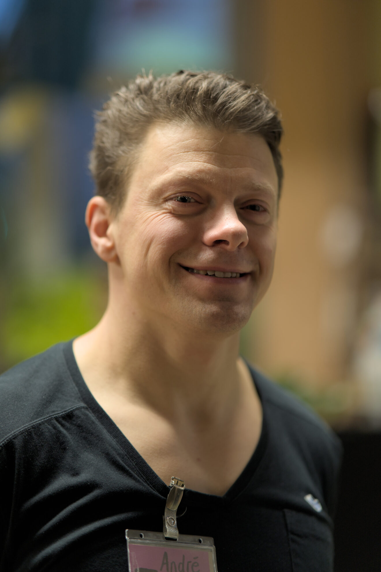 Ein Mann, seitlich fotografiert, lächelt in die Kamera. Er hat hell-braune Haare und trägt an seinem Oberteil ein Namensschild mit der Aufschrift André.