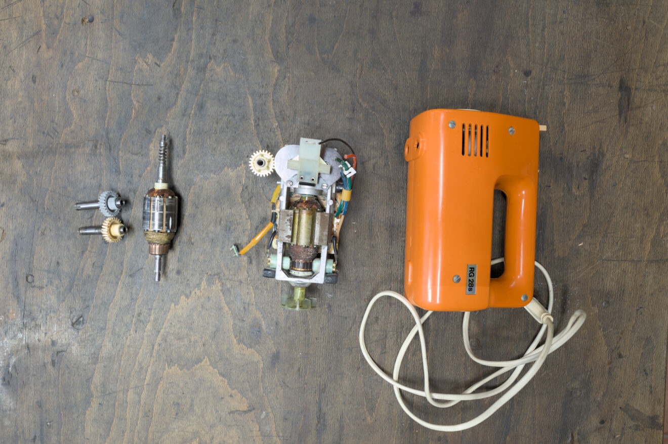 Aus der Vogelperspektive sind folgende Gegenstände auf der Werkbank zu sehen: ein orangefarbenes Rührgerät mit weißem Kabel, dem Motor und Getriebe, Anker und zwei Hohlwellen mit Zahnrädern.
