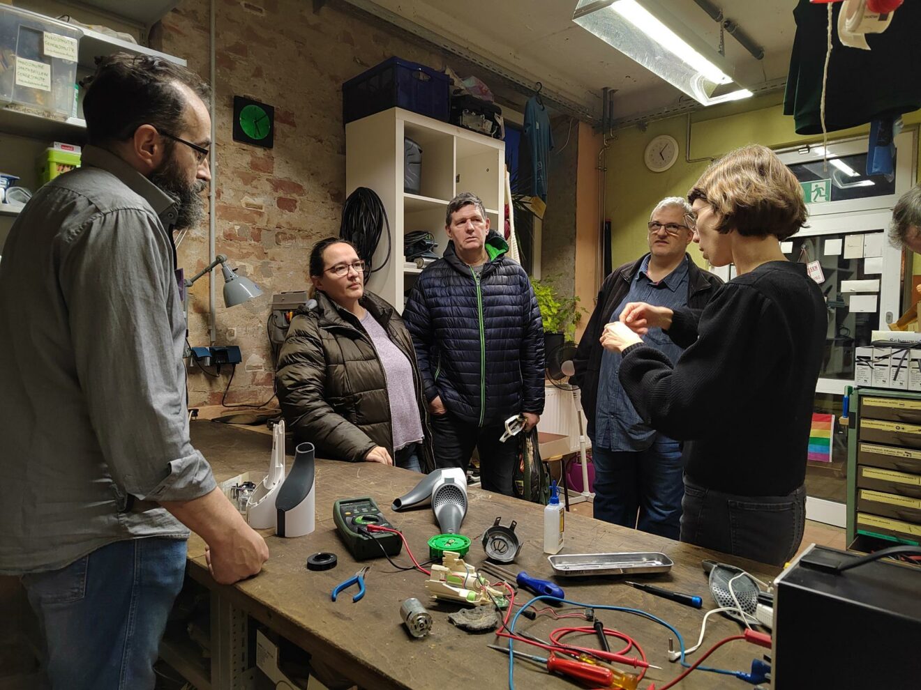 Fünf Menschen stehen im Café kaputt um eine Werkbank. Auf der Werkbank liegt ein geöffneter Handstaubsauger. Eine Dolmetscherin steht rechts und gebärdet. 