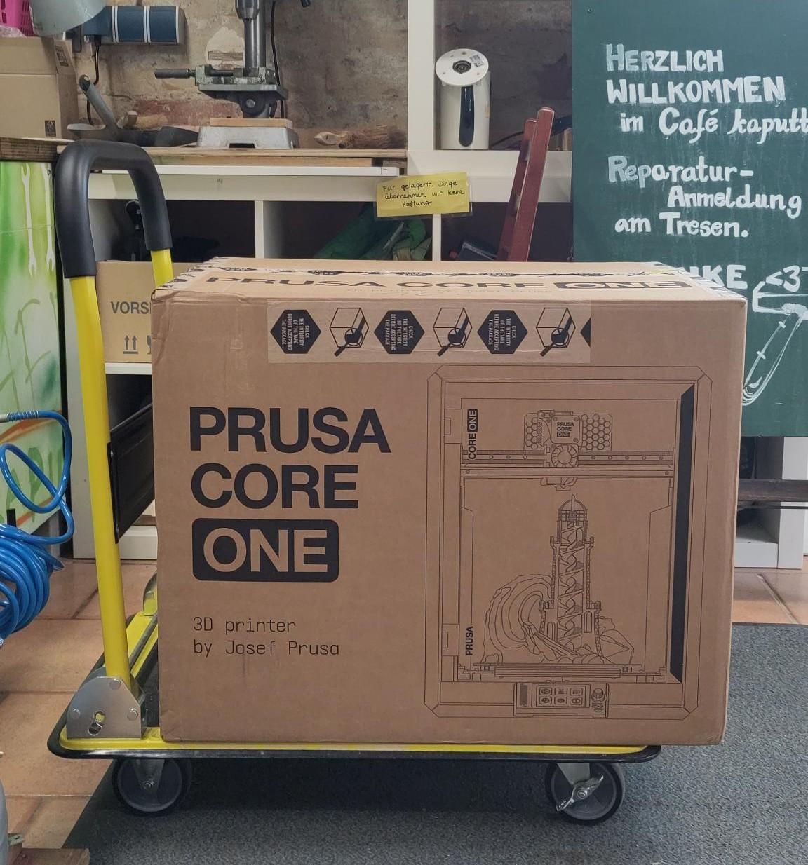 Das Paket mit unserem neuen 3D Drucker "Prusa Core One" auf einem Rollwagen vor dem Schild "Herzliche Willkommen im Café kaputt".