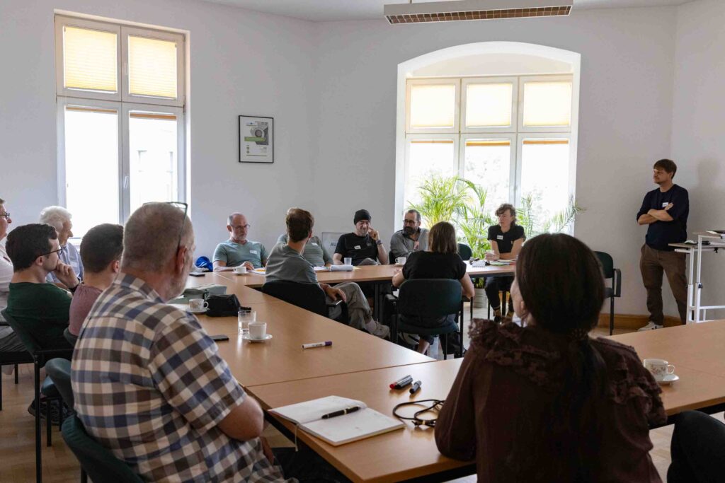 In einem Raum sitzen an Menschen an Tischen, die in einem U angeordenet sind. Vor Kopf steht ein Mensche, der einen Vortrag hält.
