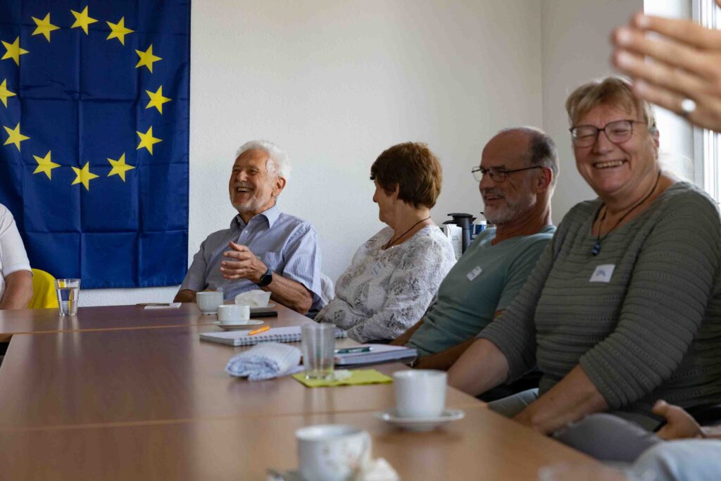 Vier ersonen an einem Tisch sitzen vor Kaffee-Tassen und lachen. Im Hintergrund ist der Ausschnitt einer Europa-Flagge zu sehen. 